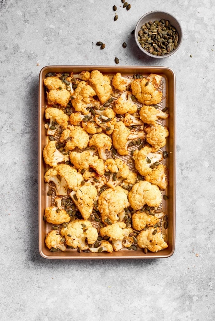 This easy roasted cauliflower is jazzed up with pepitas, cumin, smoky ancho, garlic, and lime. Crispy on the outside, tender inside! Pan of roasted cauliflower with pepitas set next to bowl of pepitas.