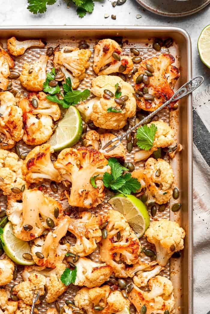 This easy roasted cauliflower is jazzed up with pepitas, cumin, smoky ancho, garlic, and lime. Crispy on the outside, tender inside! Mexican roasted cauliflower on sheet pan with pepitas, lime wedges, and cilantro.