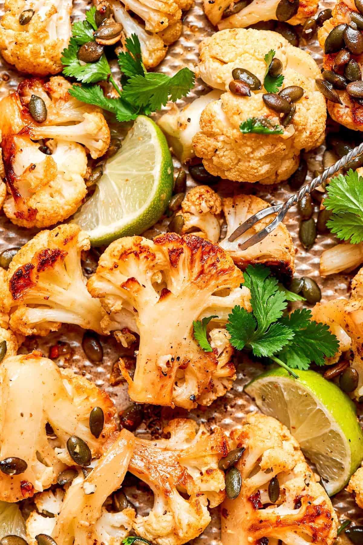 This easy roasted cauliflower is jazzed up with pepitas, cumin, smoky ancho, garlic, and lime. Crispy on the outside, tender inside! Mexican roasted cauliflower with pepitas, lime wedges, and cilantro.