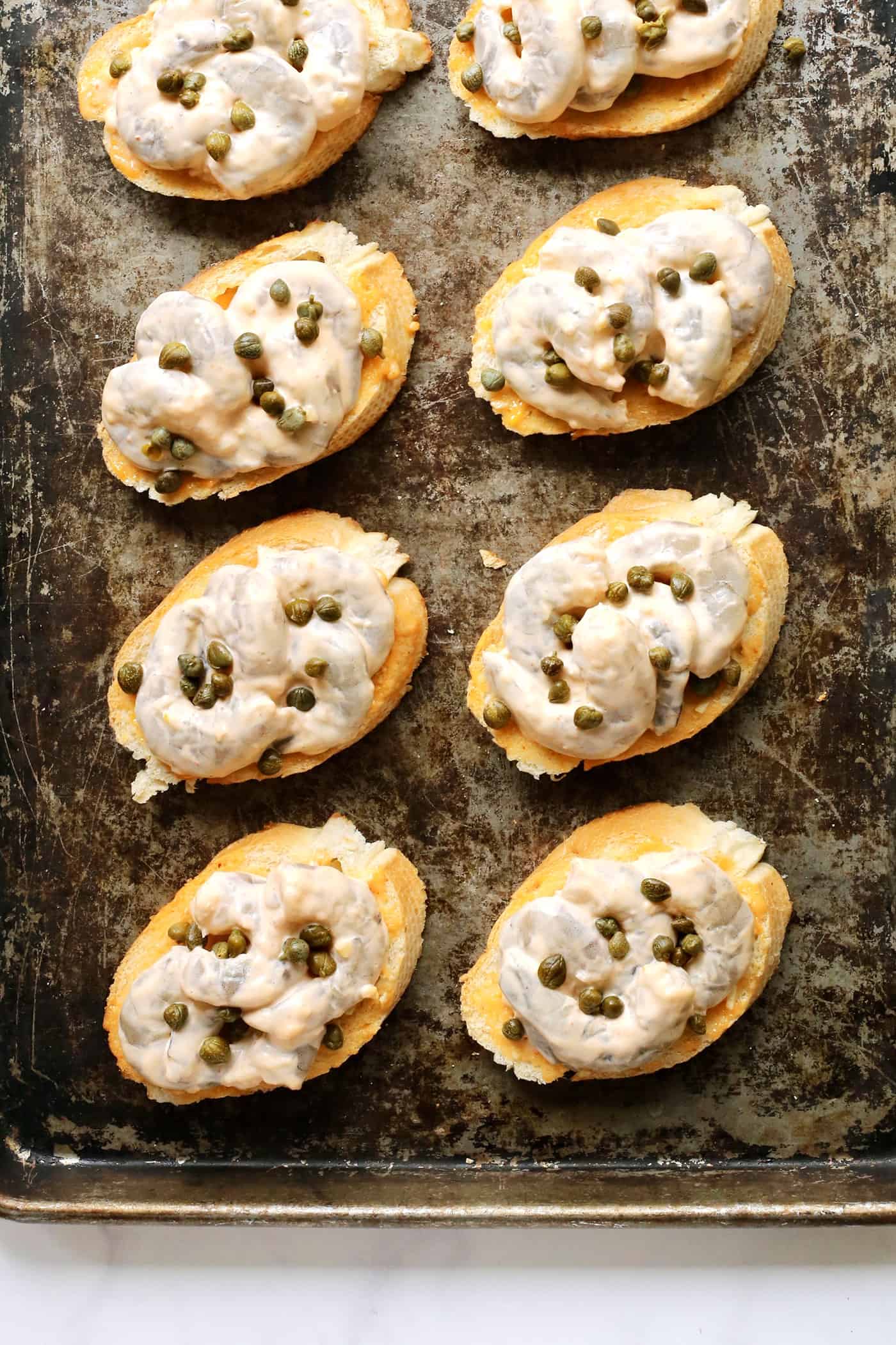 Sriracha garlic shrimp toast is made with tender shrimp, spicy sriracha mayo, and lightly crispy toast! An easy appetizer or light meal. Shrimp toast topped with capers on a baking sheet.