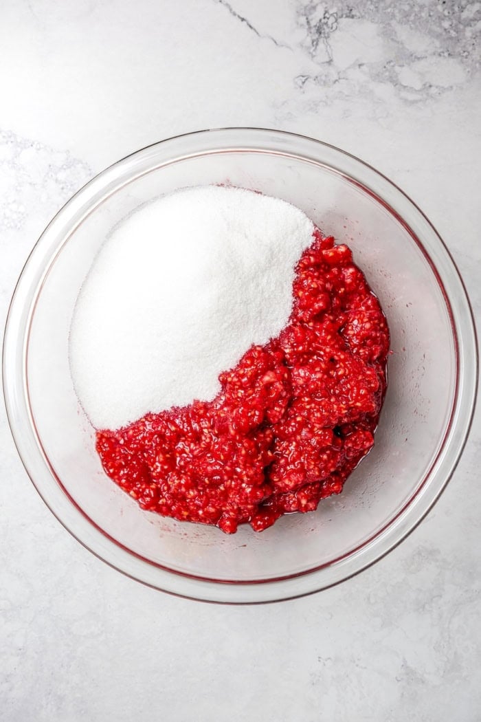 This Raspberry Freezer Jam is the easiest you'll ever make—no canning needed! It's a delicious way to extended the life of your berries. Overhead view of sugar and mashed raspberries in bowl