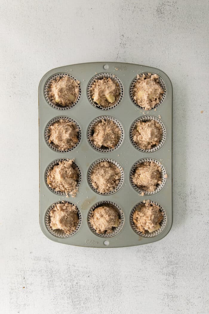 Make moist, tender apple muffins studded with juicy apple bites! Topped with cinnamon streusel, these easy muffins are great for fall baking. Apple muffins with cinnamon sugar crumble in a muffin tin ready to be baked