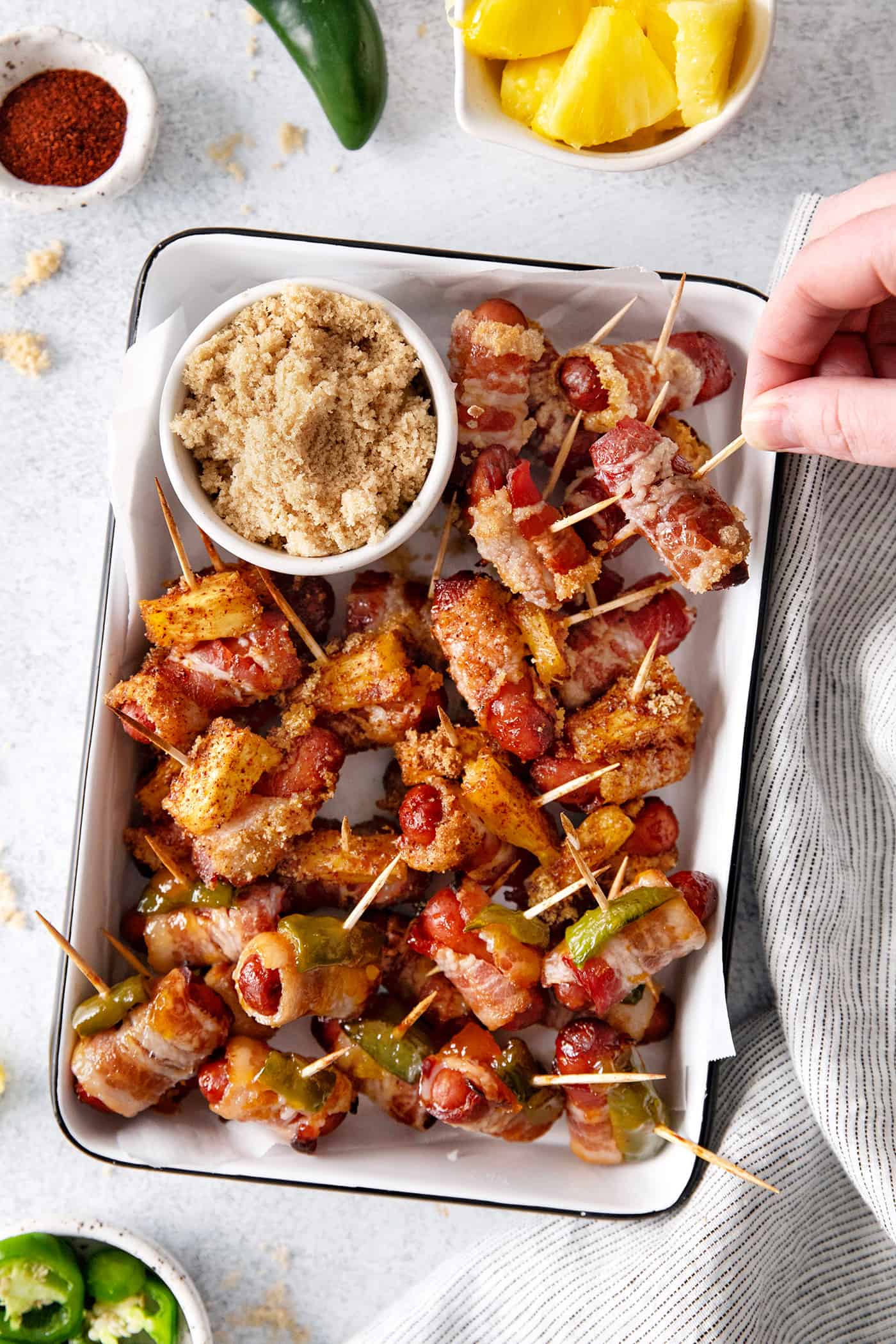 These Bacon Wrapped Smokies are the perfect game day appetizer. Switch things up by adding pineapple or jalapeno slices! A hand reach for a bacon wrapped little smokie on a serving dish