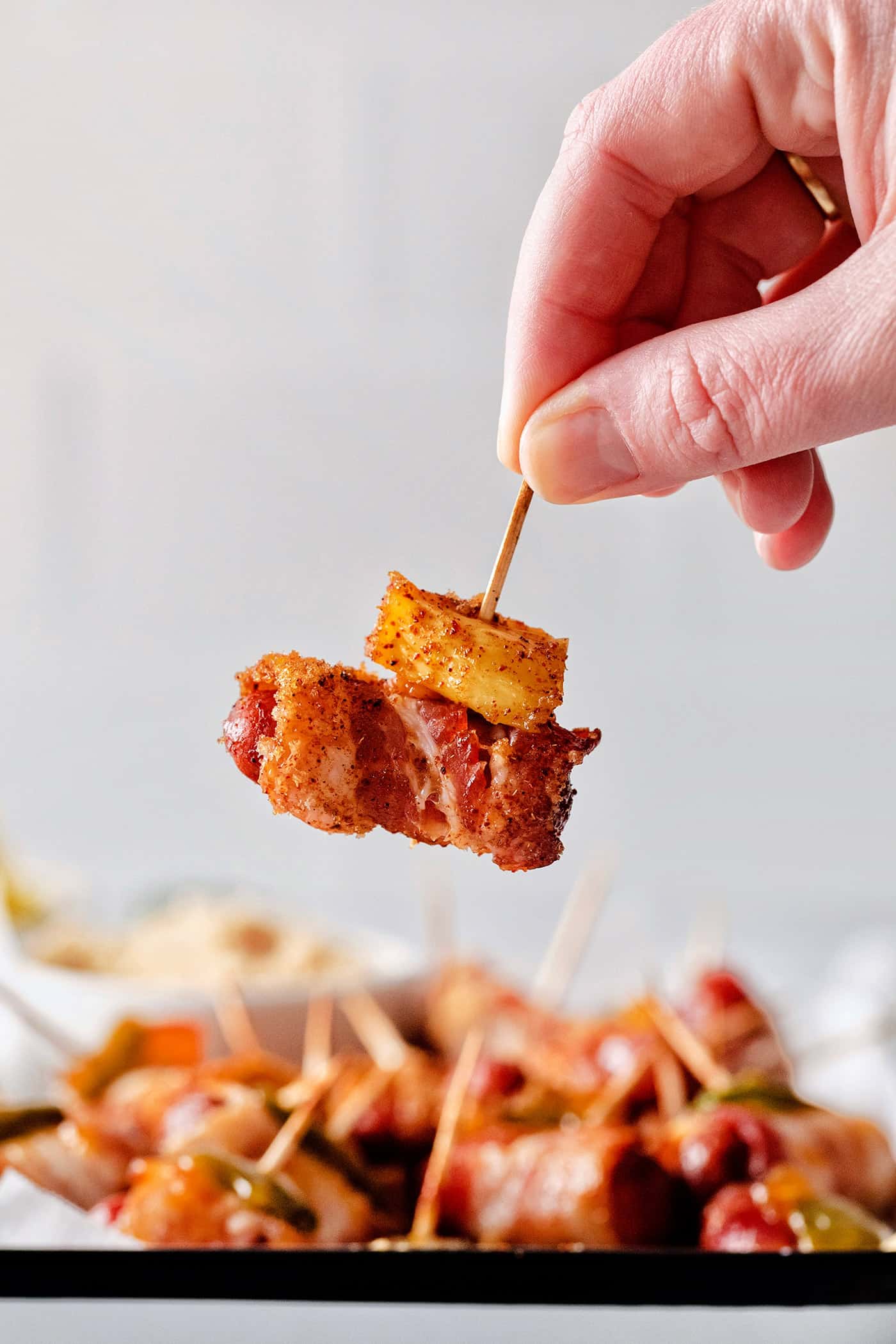 These Bacon Wrapped Smokies are the perfect game day appetizer. Switch things up by adding pineapple or jalapeno slices! A hand holding a bacon wrapper weiner with pineapple on a toothpick