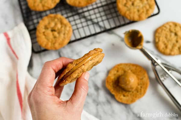 Peanut butter sandwich cookies combine soft, chewy peanut butter cookies with a rich and creamy filling, for double the flavor in every bite! Overhead view of a hand holding a peanut butter sandwich cookie with a partially assembled sandwich cookie below, next to peanut butter cookies on a wire rack.