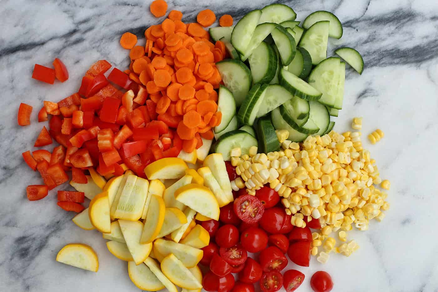 Chopped tomatoes, cucumbers, corn, peppers, carrots and squash on a countertop.