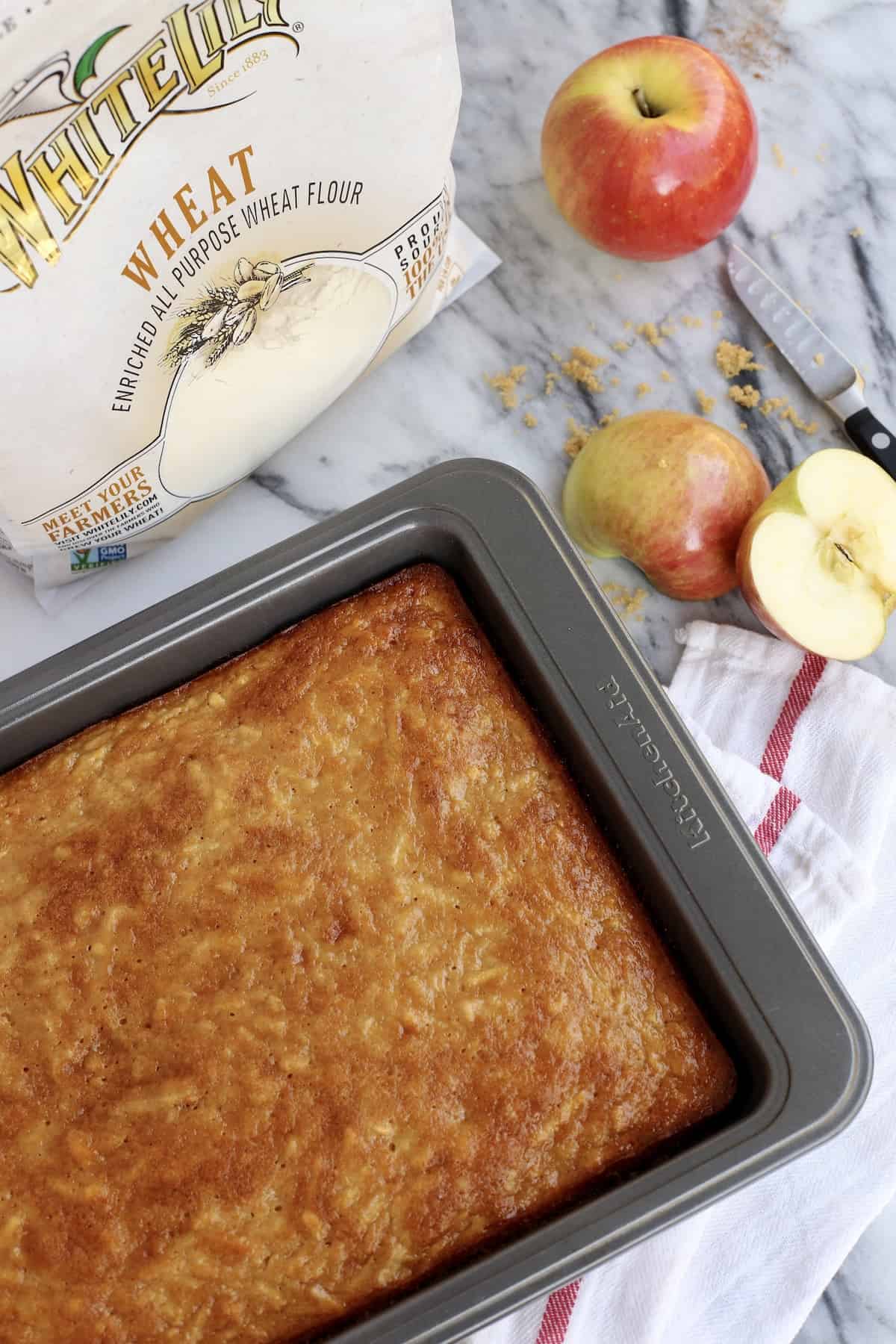 This easy apple cake is a moist, fluffy fall cake recipe filled with warm spice and fresh apples, frosted with brown sugar buttercream! Unfrosted apple cake in a pan next to a bag of flour and apples