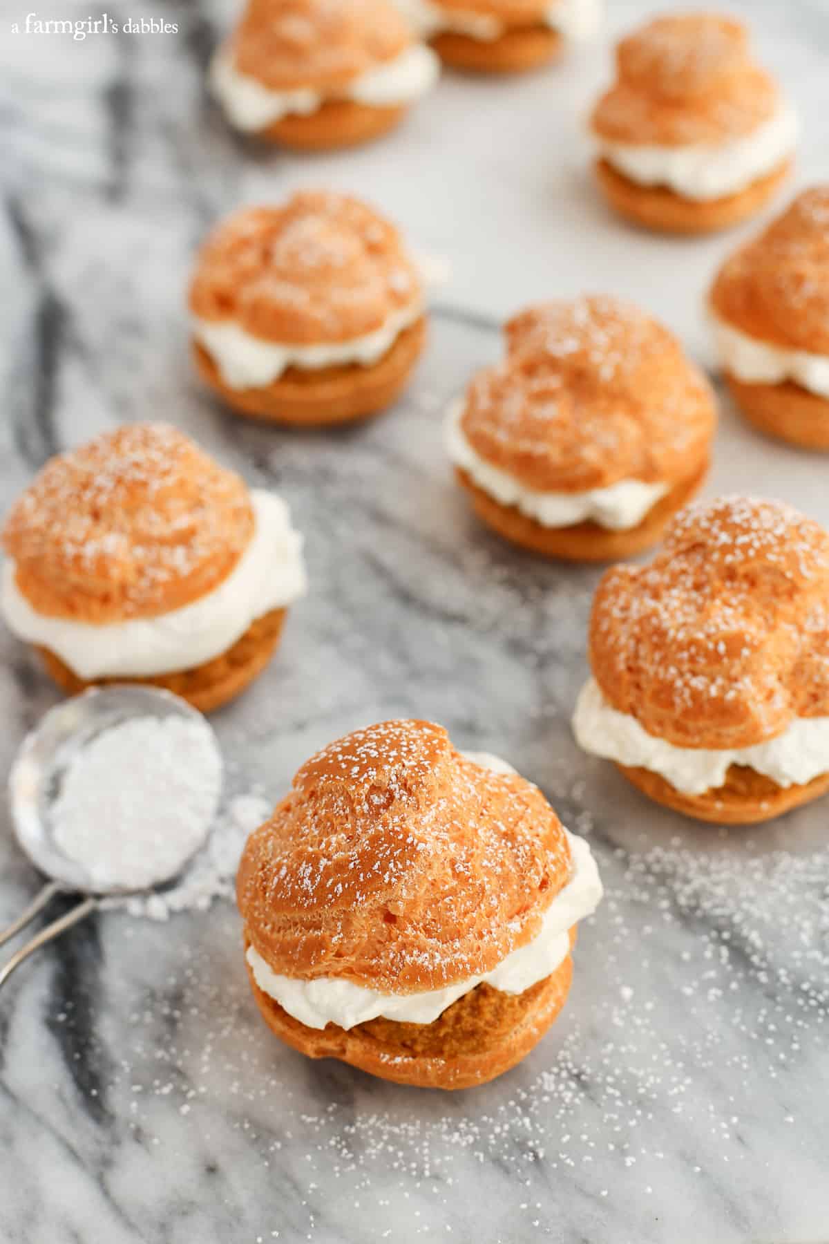 This pumpkin pie cream puff recipe is the perfect dessert for Thanksgiving. They're layered with pumpkin pie filling and maple whipped cream! Pumpkin Pie Cream Puffs on a marble table sprinkled with powered sugar.
