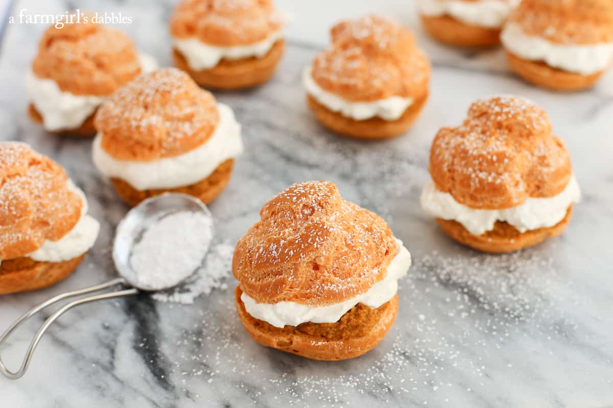 This pumpkin pie cream puff recipe is the perfect dessert for Thanksgiving. They're layered with pumpkin pie filling and maple whipped cream! Homemade Cream Puffs on a table with a spoon of powdered sugar.
