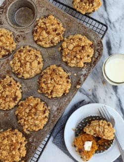 Overhead view of pumpkin muffins topped with streusel in a muffin tin next to one muffin cut in half on a plate.