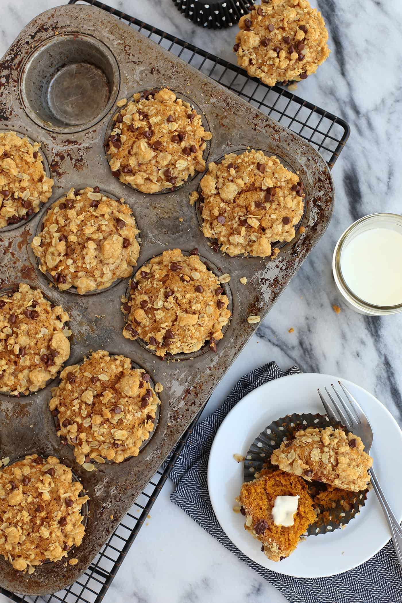 Super moist and perfectly spiced, this pumpkin muffins recipe features a crispy, buttery oat streusel dotted with melty chocolate chips. Overhead view of pumpkin muffins topped with streusel in a muffin tin next to one muffin cut in half on a plate.