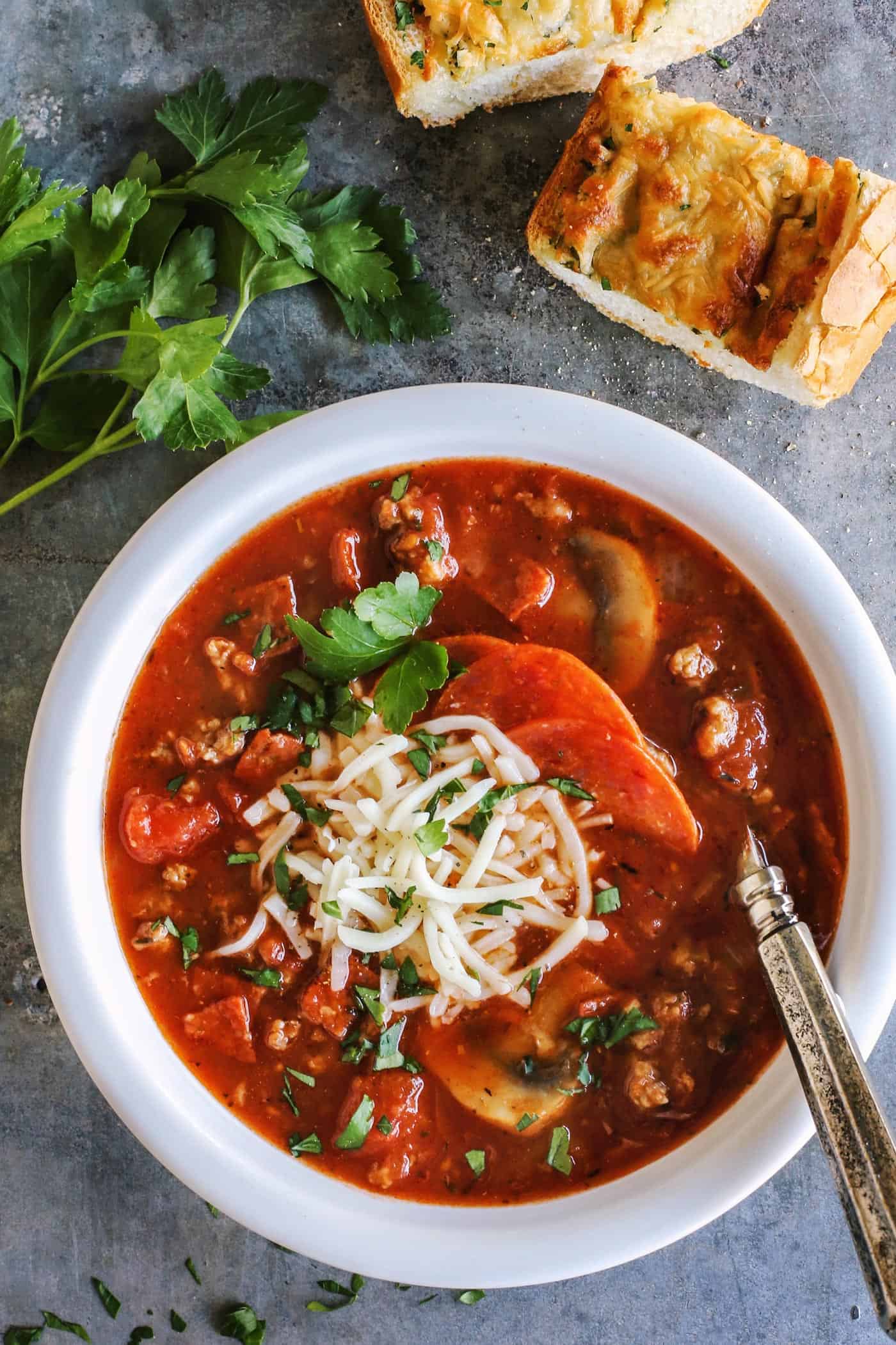 Overhead view of hearty pizza soup in a bowl topped with shredded cheese, next to slices of garlic bread.