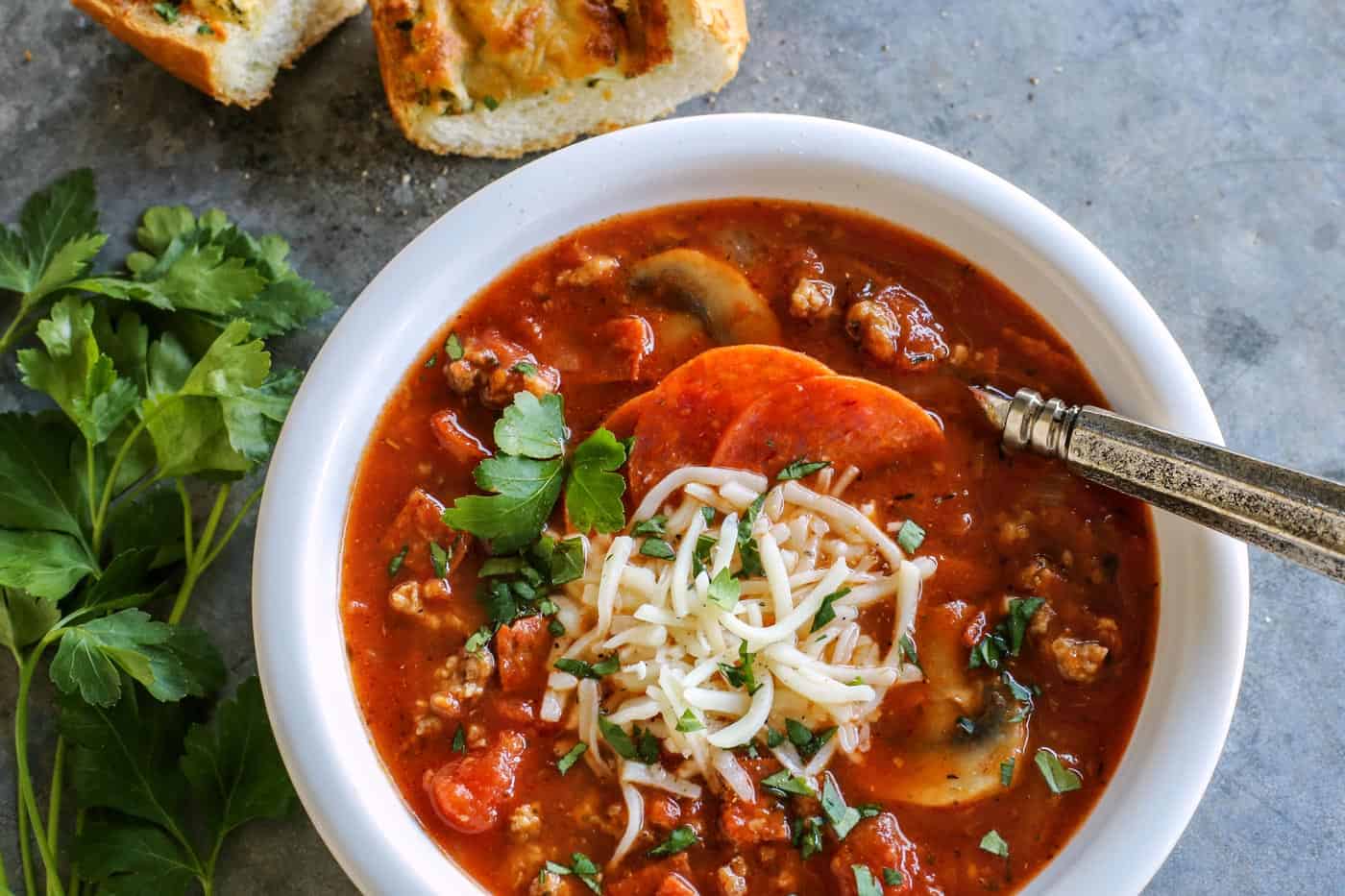 Overhead view of pizza soup with sausage and mushrooms in a bowl topped with shredded cheese, next to slices of garlic bread.