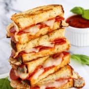 A stack of pizza grilled cheese halves with basil leaves and pizza sauce on the side for dipping.