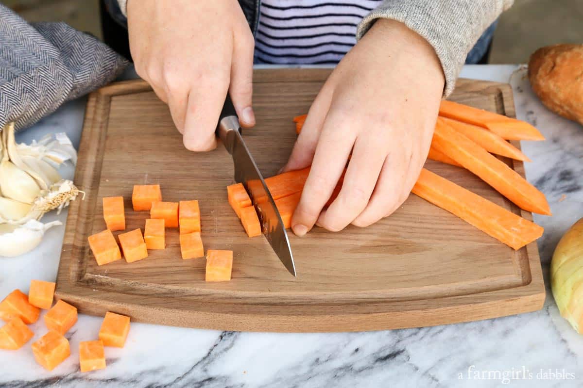 This Southwestern slow cooker beef stew with sweet potatoes combines hearty ingredients in a rich broth, brightened with a squeeze of lime! Hands using a large knife to cut sweet potatoes into cubes on a wooden cutting board.