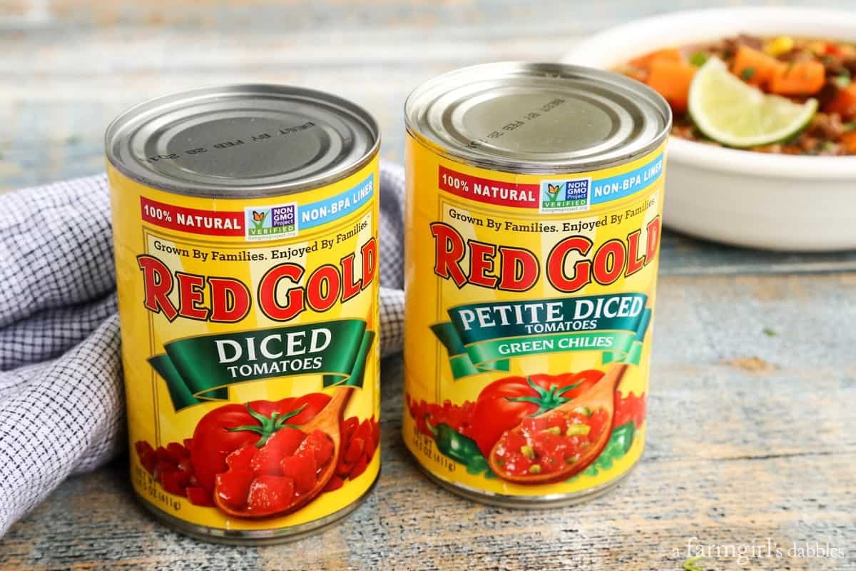 This Southwestern slow cooker beef stew with sweet potatoes combines hearty ingredients in a rich broth, brightened with a squeeze of lime! Two cans of Red Gold tomatoes on a countertop with a bowl of stew in the background.