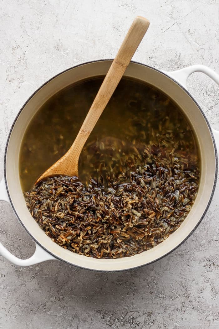 This creamy Wild Rice Casserole with sausage and mushrooms is deeply savory, hearty, and perfect for cozy dinners and holiday celebrations! Wooden spoon stirring wild rice in pot of broth.