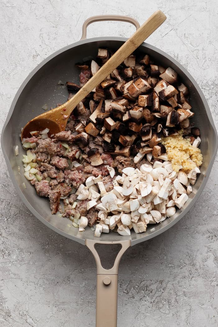 This creamy Wild Rice Casserole with sausage and mushrooms is deeply savory, hearty, and perfect for cozy dinners and holiday celebrations! Wooden spoon stirring mushrooms and garlic into pan of browned sausage.