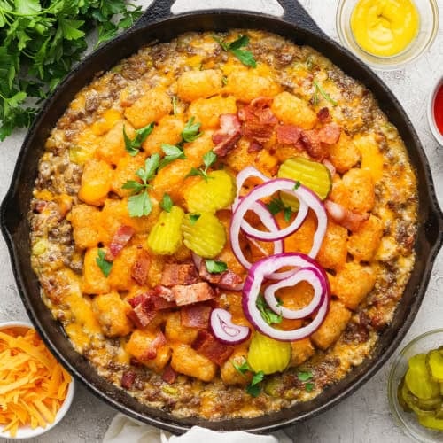 Bacon cheeseburger tater tot casserole in skillet with pickles and onions on top.
