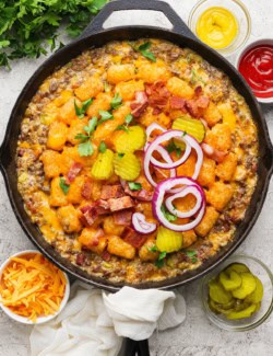 Bacon cheeseburger tater tot casserole in skillet with pickles and onions on top.