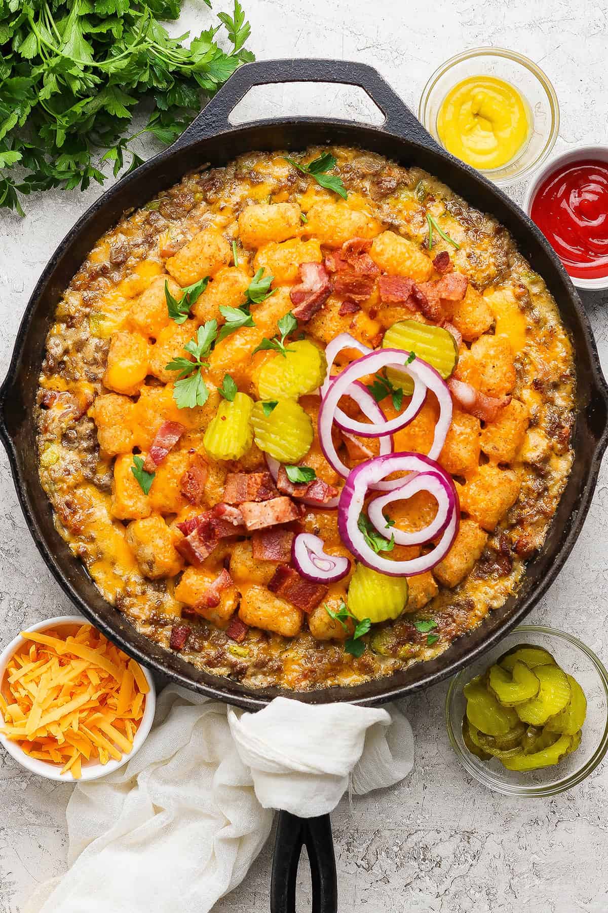 Crispy tater tots, ground beef, smoky bacon and cheese make this bacon cheeseburger tater tot casserole taste like a burger in hot dish form! Bacon cheeseburger tater tot casserole in skillet with pickles and onions on top.