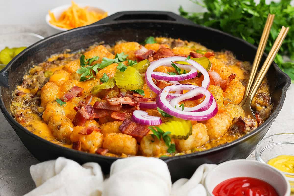 Crispy tater tots, ground beef, smoky bacon and cheese make this bacon cheeseburger tater tot casserole taste like a burger in hot dish form! Skillet of bacon cheeseburger casserole with two spoons.