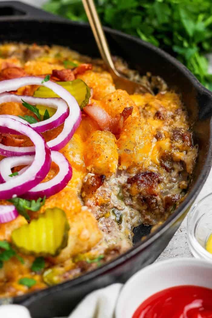 Crispy tater tots, ground beef, smoky bacon and cheese make this bacon cheeseburger tater tot casserole taste like a burger in hot dish form! Spoon in skillet of bacon cheeseburger casserole.