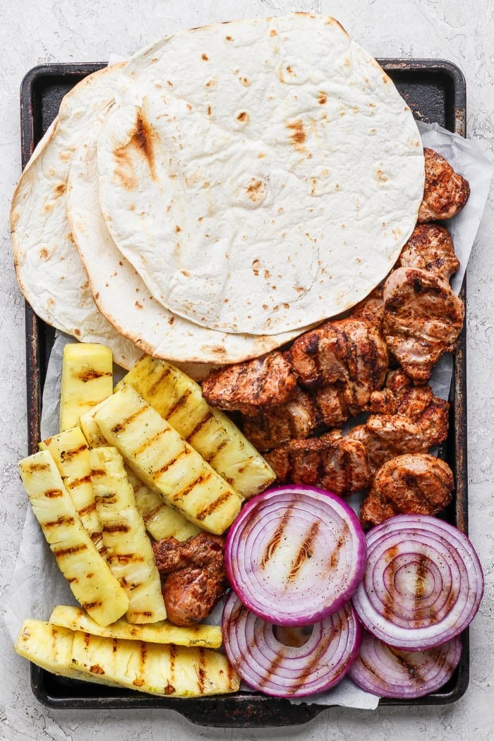 This Grilled Pork and Pineapple Tacos recipe a much easier twist on tacos al pastor. It promises to be your next taco night hit! Overhead view of flour tortillas, grilled pork, pineapple, and red onion slices on sheet pan