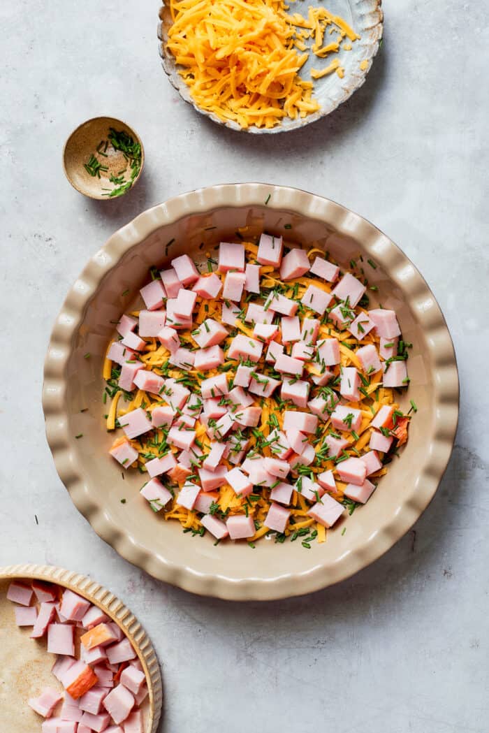 This cheesy crustless quiche recipe with ham, tangy Dijon, and chives is an easy baked dish for the holidays. Assembling the layers of cheese, chives, and ham for the quiche.
