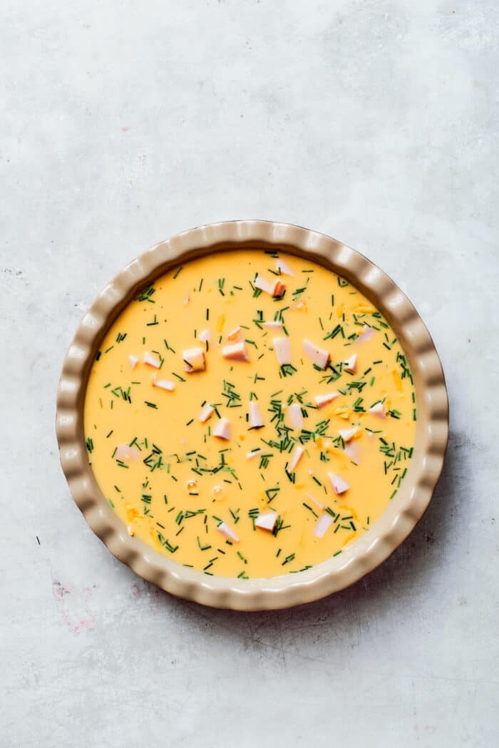 This cheesy crustless quiche recipe with ham, tangy Dijon, and chives is an easy baked dish for the holidays. Pouring the egg mixture over the cheese and ham layers.