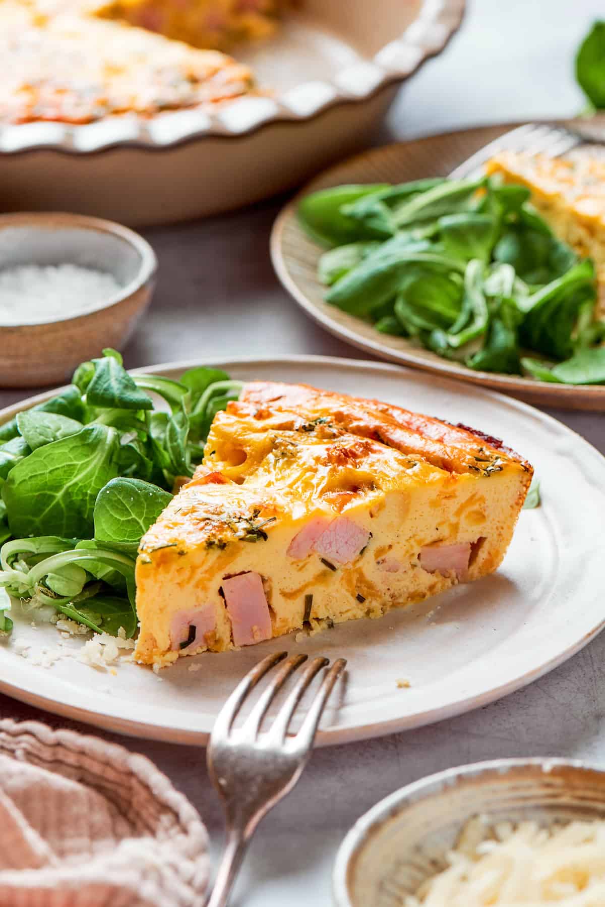 This cheesy crustless quiche recipe with ham, tangy Dijon, and chives is an easy baked dish for the holidays. Close-up of a slice of creamy ham quiche on a serving plate with salad on the side.