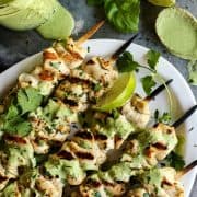 Overhead view of grilled chicken skewers with jar of green curry yogurt sauce and lime wedges