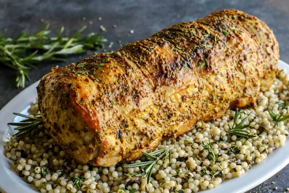 This pork loin roast recipe is juicy and tender, slathered with a savory mustard mixture and oven-roasted for a satisfying, easy pork dinner. A whole pork loin roast served over a bed of pearl couscous on a plate.