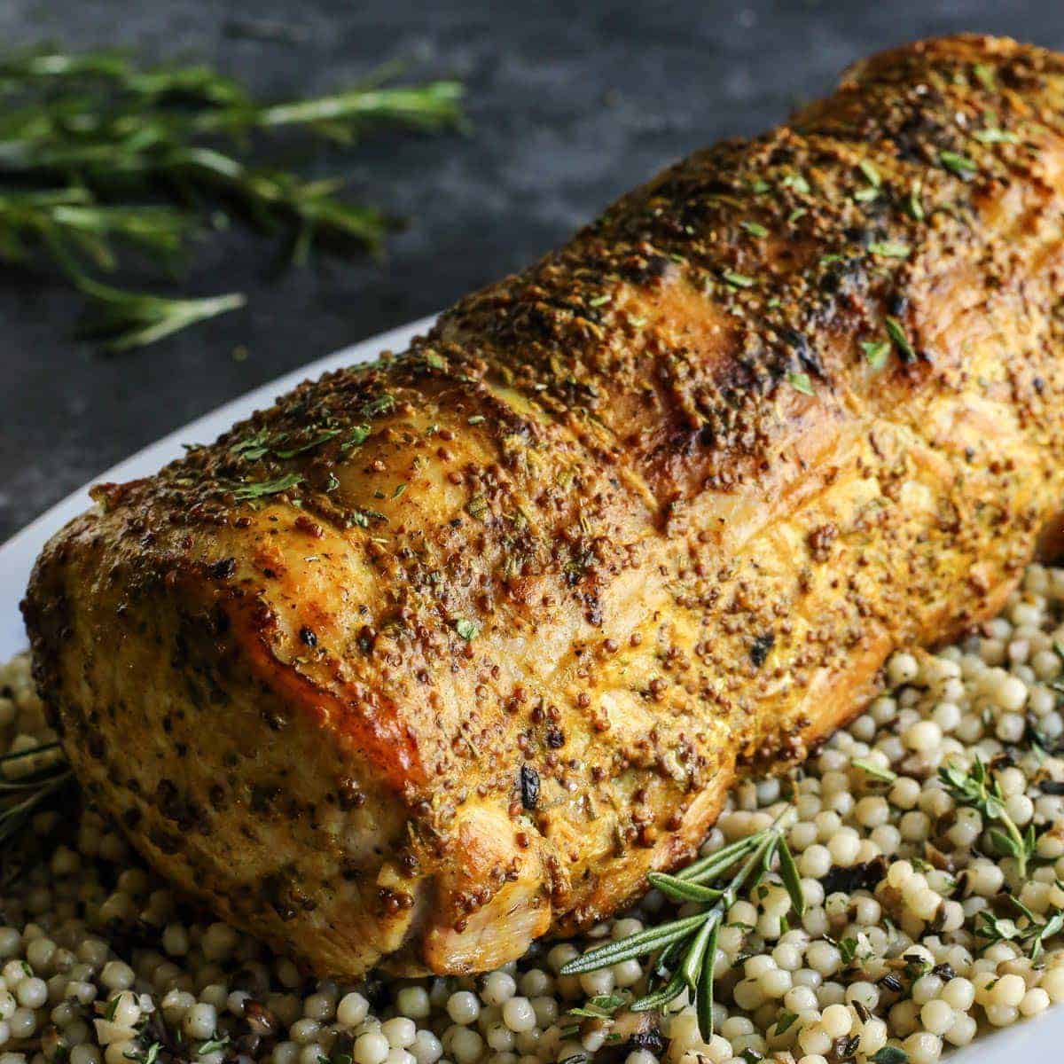This pork loin roast recipe is juicy and tender, slathered with a savory mustard mixture and oven-roasted for a satisfying, easy pork dinner. A whole pork loin roast served over a bed of pearl couscous on a plate.