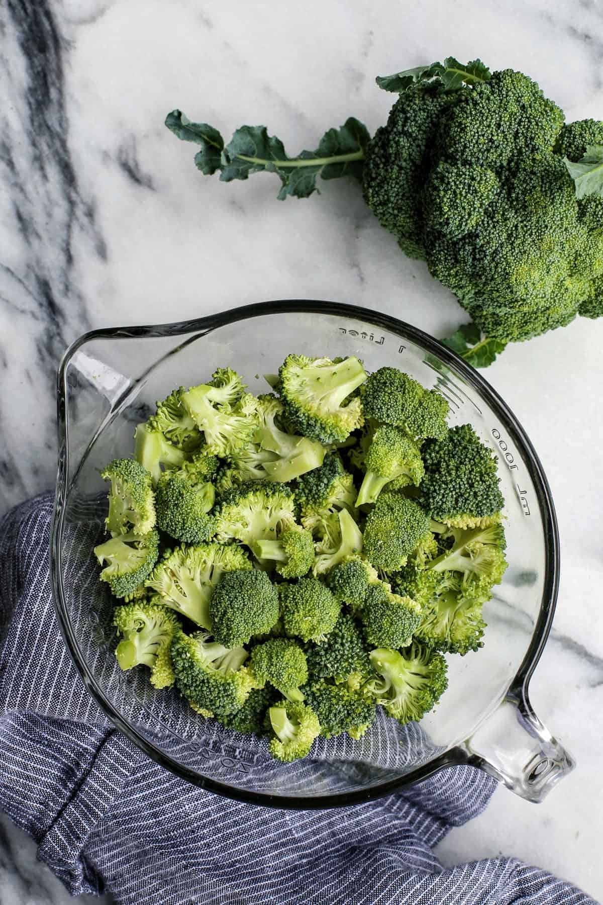 Extra cheesy and creamy, this broccoli cheddar soup recipe is loaded with all kinds of wholesome veggies. Don't forget a sprinkle of bacon! Overhead view of broccoli florets in a glass bowl next to a head of broccoli on a countertop.