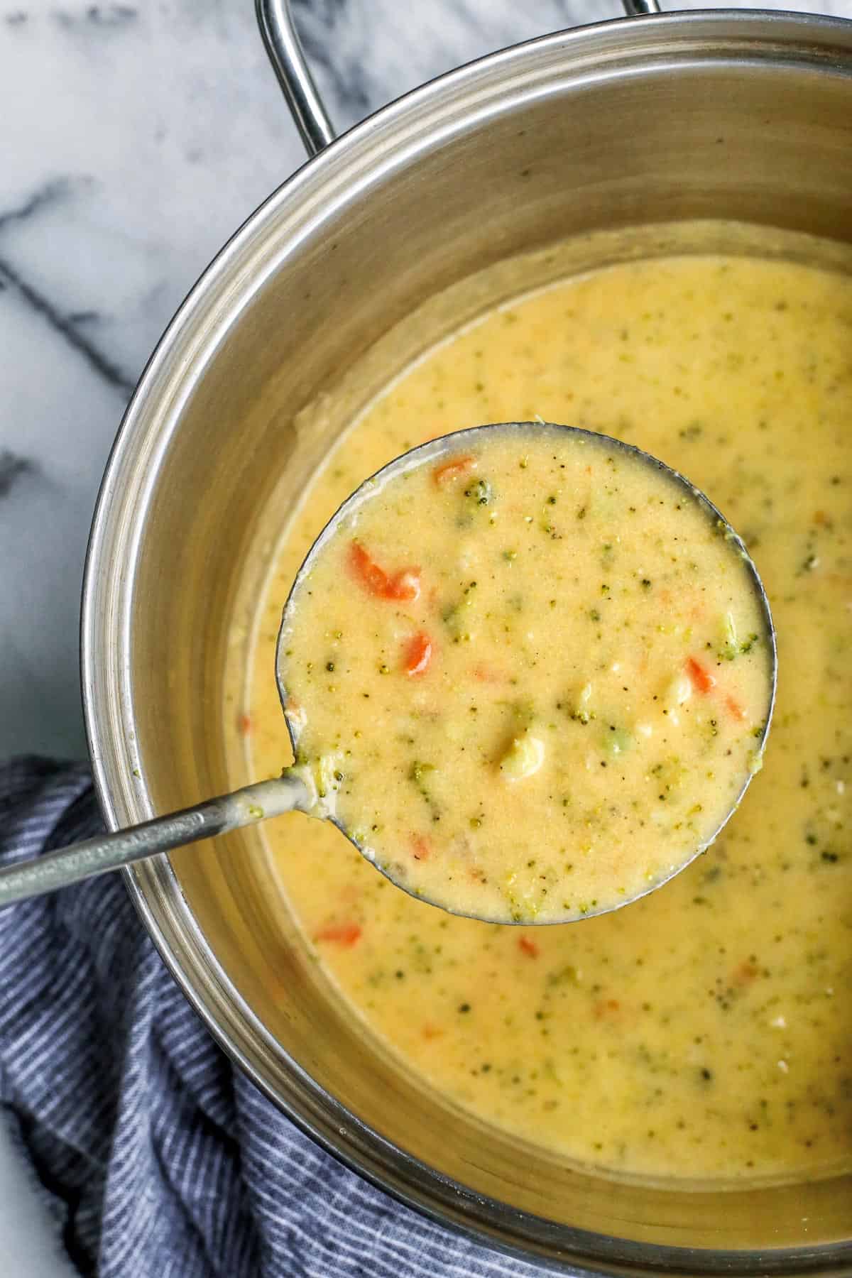 Extra cheesy and creamy, this broccoli cheddar soup recipe is loaded with all kinds of wholesome veggies. Don't forget a sprinkle of bacon! Overhead view of a ladle of broccoli cheddar soup held over the rest of the soup in a large pot.