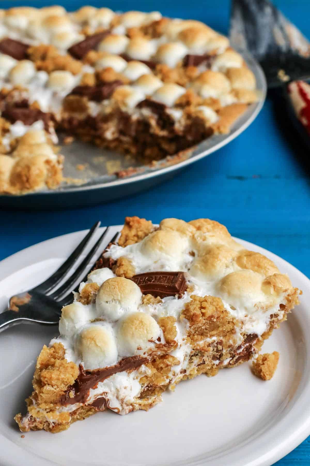 Gooey, chocolatey, and utterly delicious, this S'mores Pie allows you to enjoy the flavors of the classic summertime treat any time of the year. No campfire required! S'mores Pie in the background with a slice on a plate in front