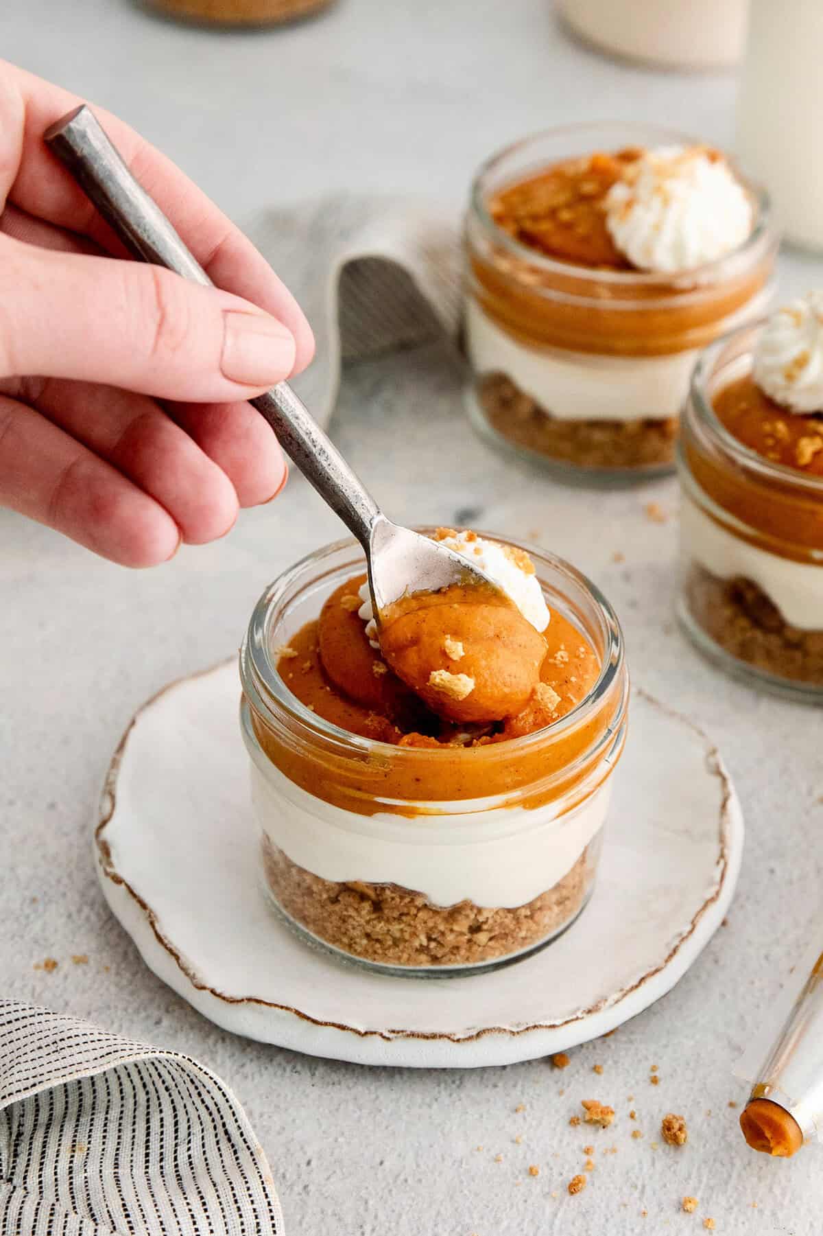 This no-bake pumpkin pie in a jar recipe with warm spices, tangy cheesecake, spiced pumpkin, and whipped cream is great for the holidays. Digging into the pumpkin pie in a jar with a spoon.