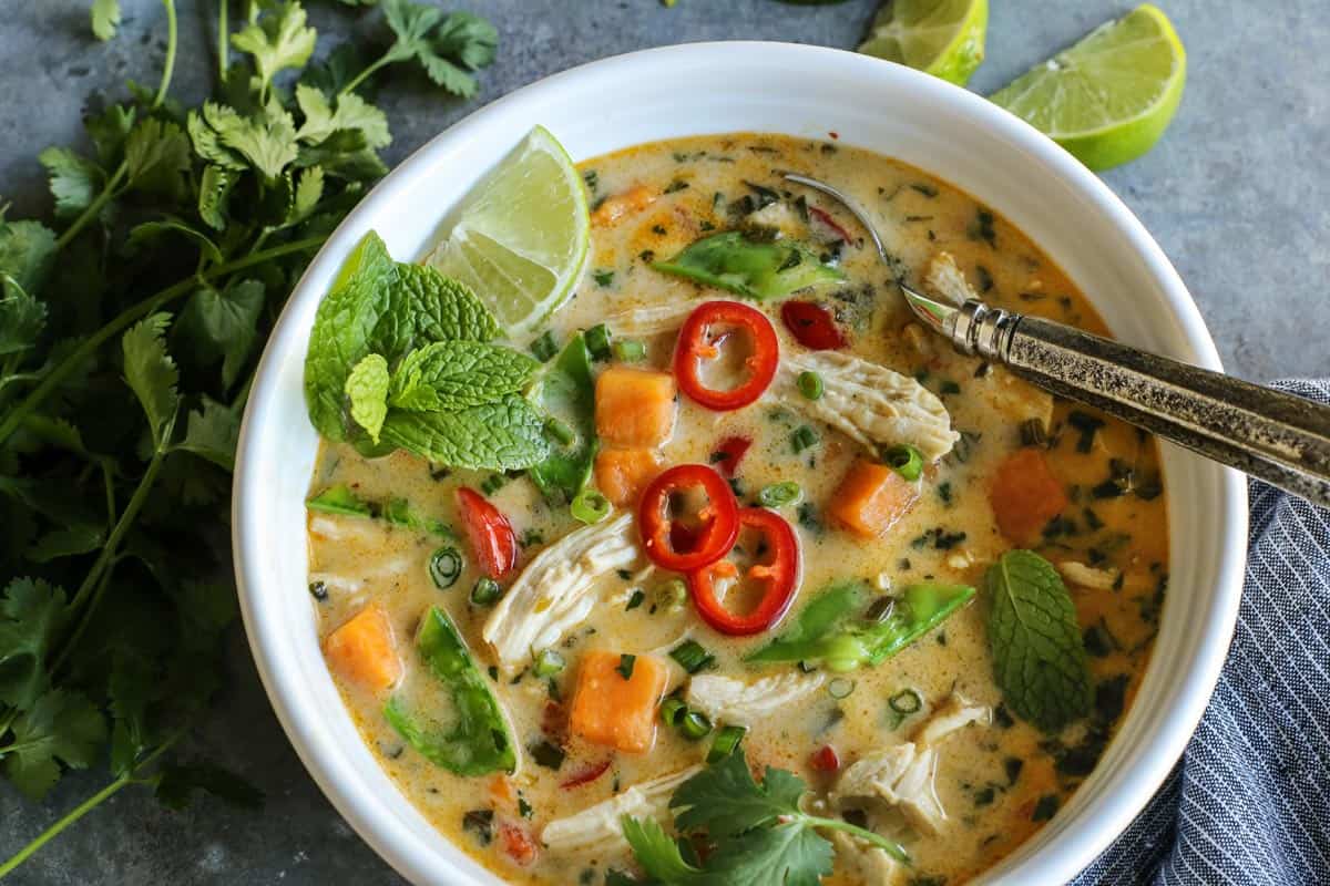With shredded chicken, chunks of sweet potato, and creamy coconut milk, this warming Thai chicken soup comes together in under an hour! Overhead view of Thai Chicken Soup in a white bowl garnished with fresh mint and sliced chilies.