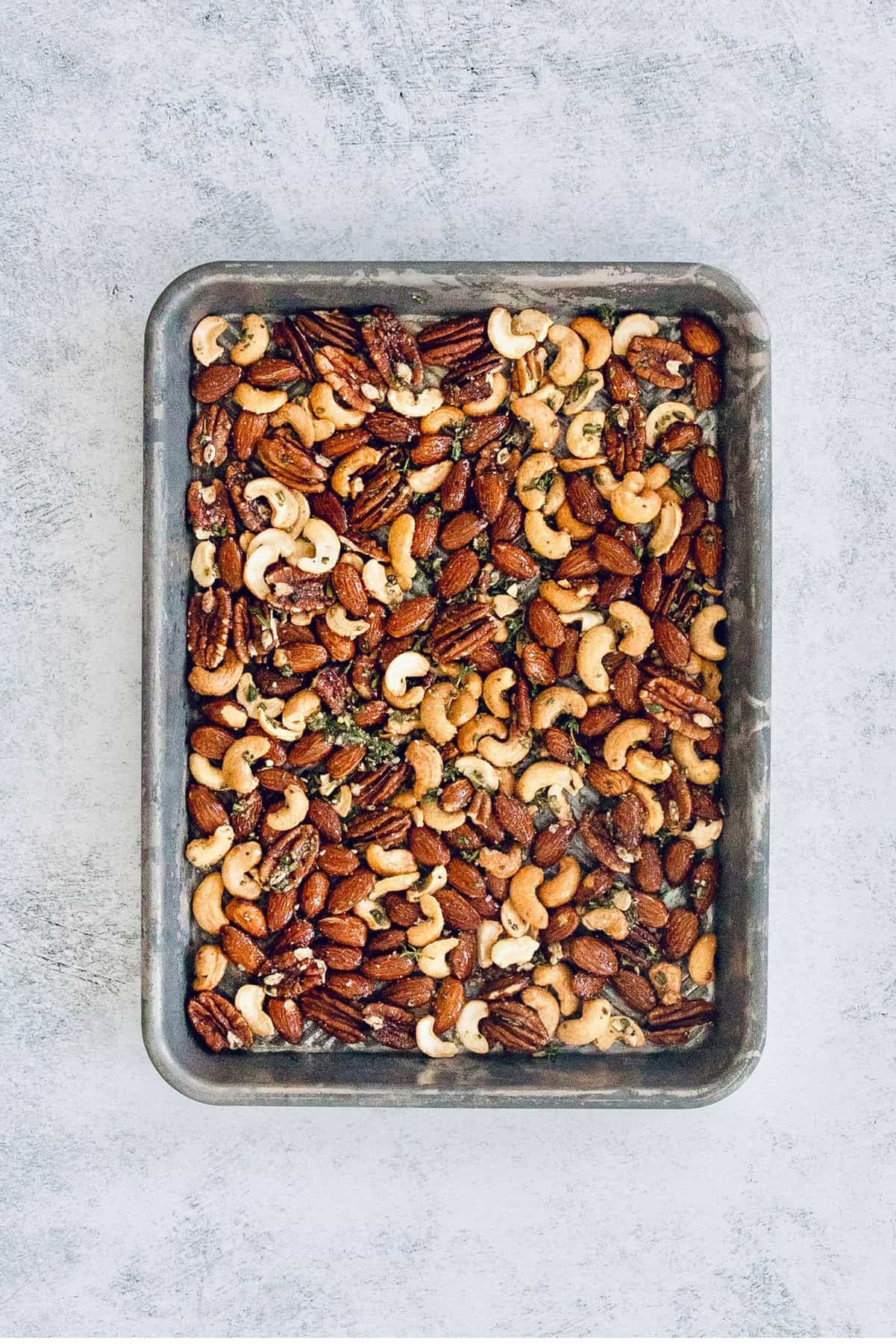 This homemade, baked spiced nuts recipe with herb-infused olive oil, cumin, spicy cayenne, and sugar is an easy holiday appetizer. Adding the seasoned nuts to the baking tray.