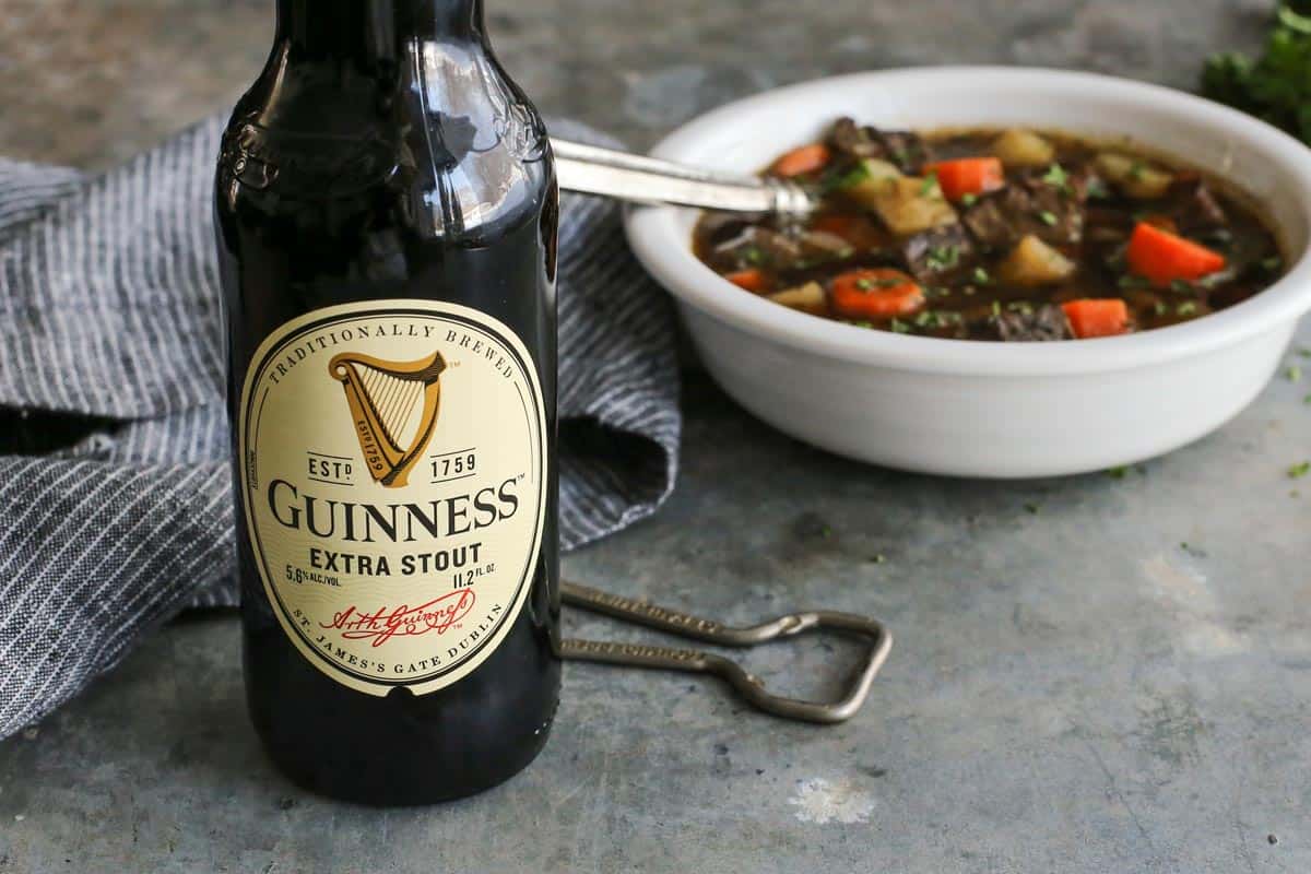 This traditional Irish beef stew is extra hearty, with tender beef and vegetables simmered in a rich beef broth with Irish Guinness beer. Guinness Extra Stout with a bowl of Irish beef stew in the background.