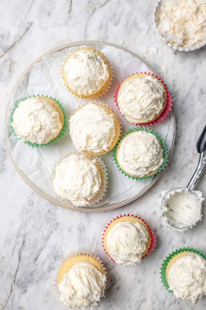 These coconut cupcakes are moist and full of coconut flavor — and just wait until you sink your teeth into the coconut buttercream frosting! Overhead view of coconut cupcakes on plate and marble countertop
