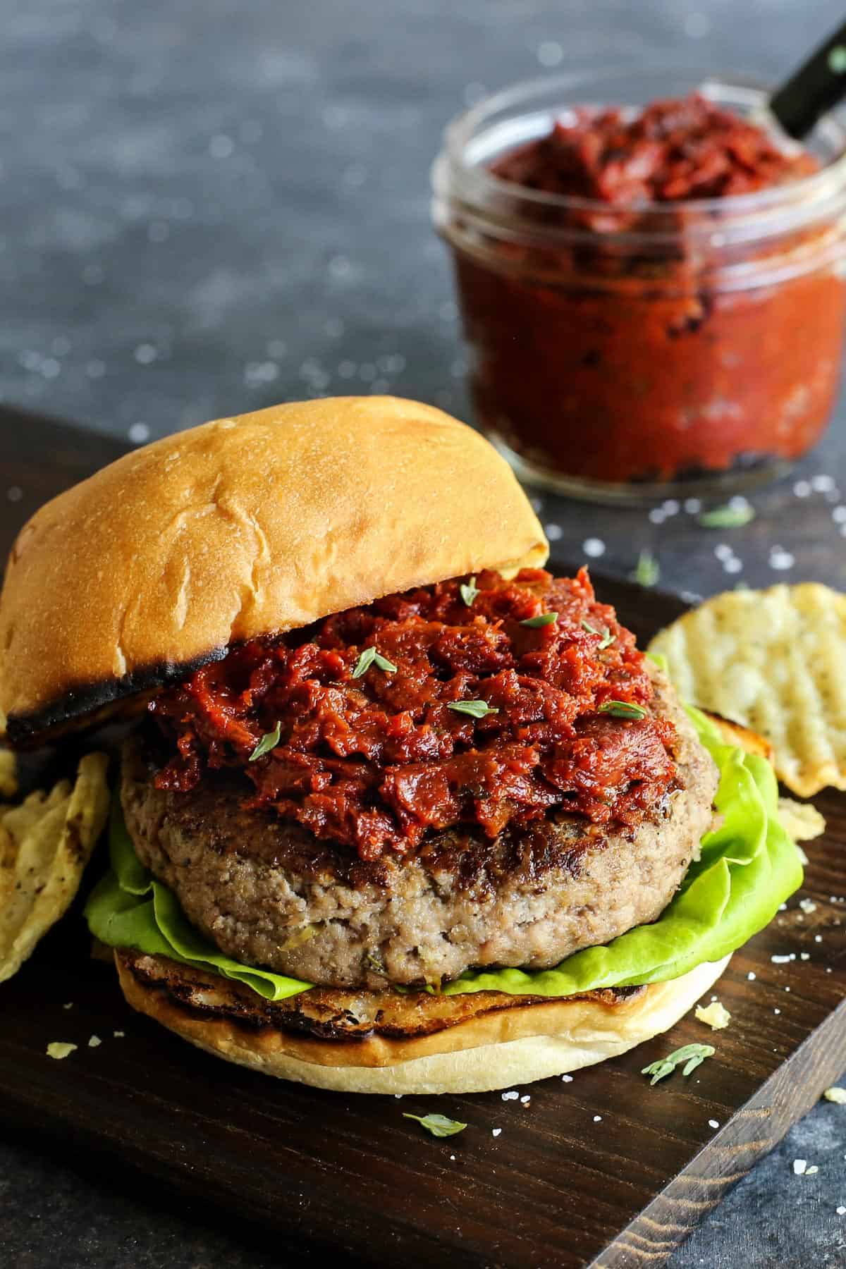 Ground Turkey Burgers feature juicy turkey patties stuffed with creamy, tangy goat cheese plus lots of fresh lemon and herbs. A ground turkey burger is served topped with sundried tomato spread with a jar of spread in the background.