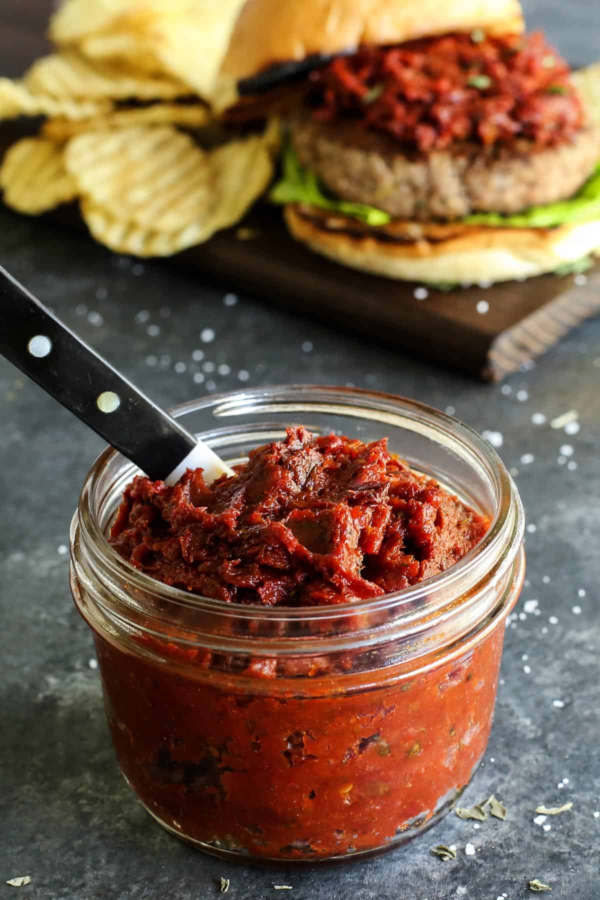 Ground Turkey Burgers feature juicy turkey patties stuffed with creamy, tangy goat cheese plus lots of fresh lemon and herbs. A jar of sun-dried tomato spread with a knife sticking out of it with a turke burger served in the background.