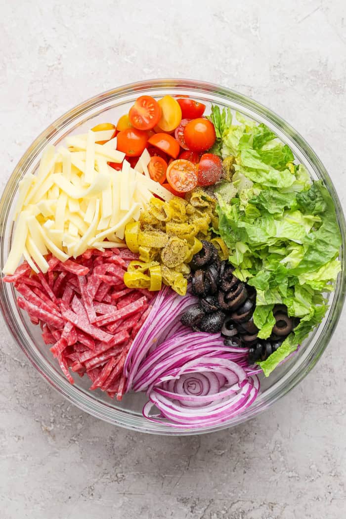 This Italian Chopped Salad is loaded with fresh veggies, salami, provolone, pepperoncini, and olives for a flavorful, satisfying meal! Overhead view of Italian chopped salad ingredients in bowl before tossing