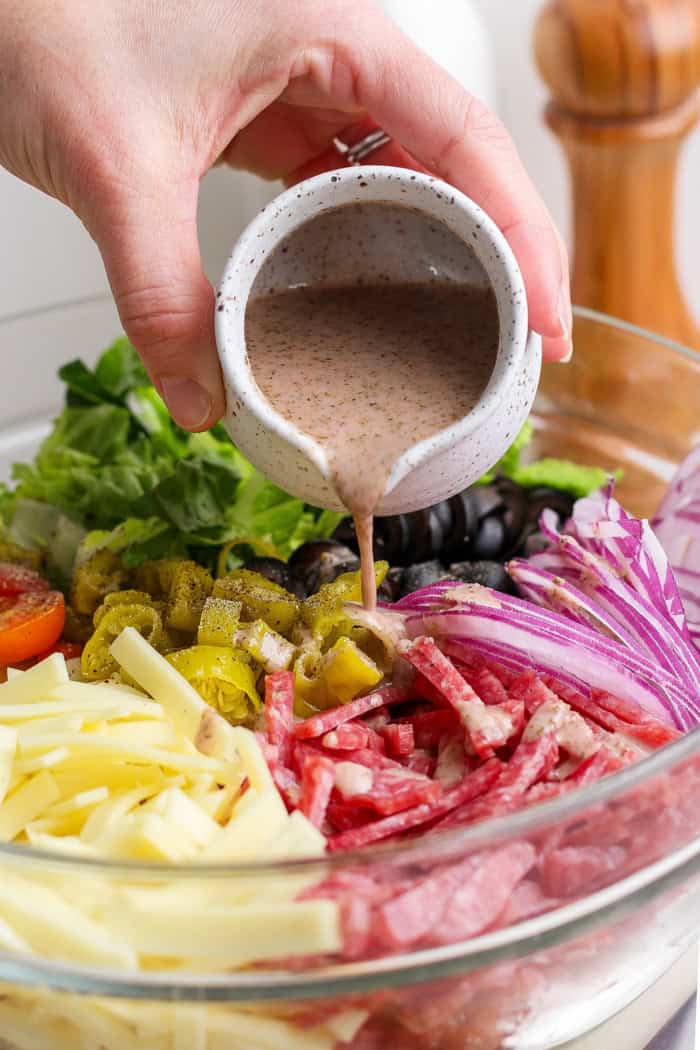 This Italian Chopped Salad is loaded with fresh veggies, salami, provolone, pepperoncini, and olives for a flavorful, satisfying meal! Pouring Italian vinaigrette into bowl of ingredients for Italian chopped salad