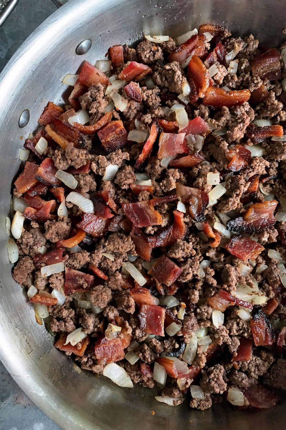 Made with bacon, ground beef, and baked beans, these easy Cowboy Beans are a hearty and flavorful side dish. They're the perfect accompaniment to anything off the grill and ideal for casual gatherings and potlucks too! skillet of browned ground beef, bacon pieces, and chopped onion