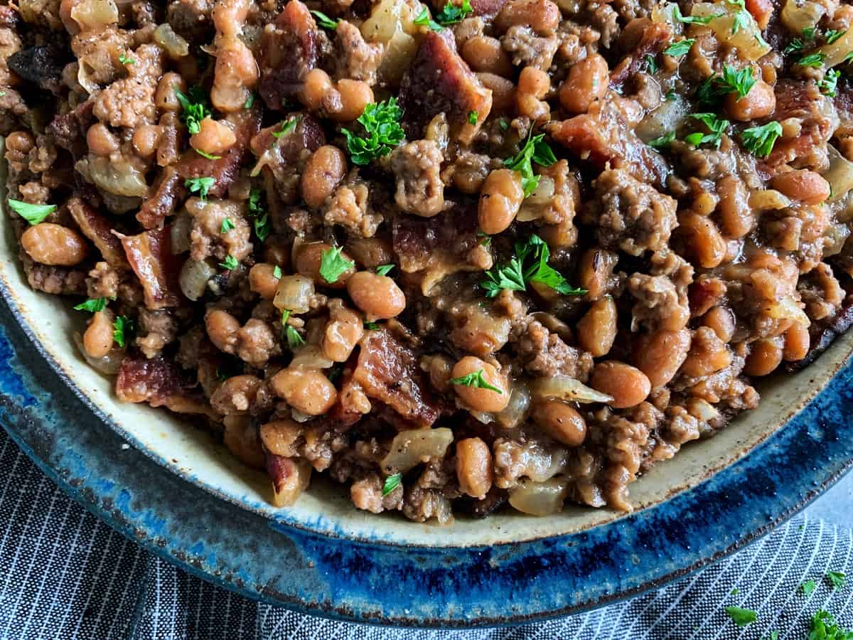 Made with bacon, ground beef, and baked beans, these easy Cowboy Beans are a hearty and flavorful side dish. They're the perfect accompaniment to anything off the grill and ideal for casual gatherings and potlucks too! dish of baked beans with bacon and ground beef