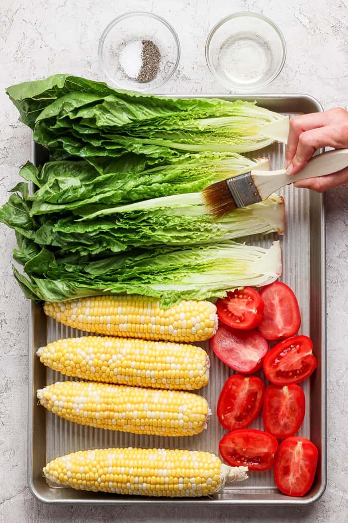 This smoky Grilled Romaine Salad is simple and summery, with tomatoes, sweet corn, crispy bacon and homemade buttermilk ranch dressing. Yum! Brushing romaine with oil, on pan with 4 ears of corn and halved roma tomatoes