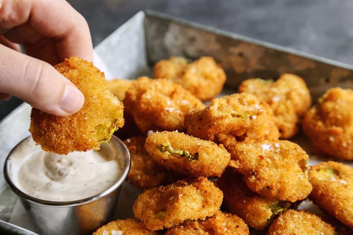 Enjoy your favorite fair food at home with these easy Fried Pickles. Dill pickles are dipped in a perfectly seasoned batter and quickly fried. a hand dipping a fried pickle slice into ranch dip